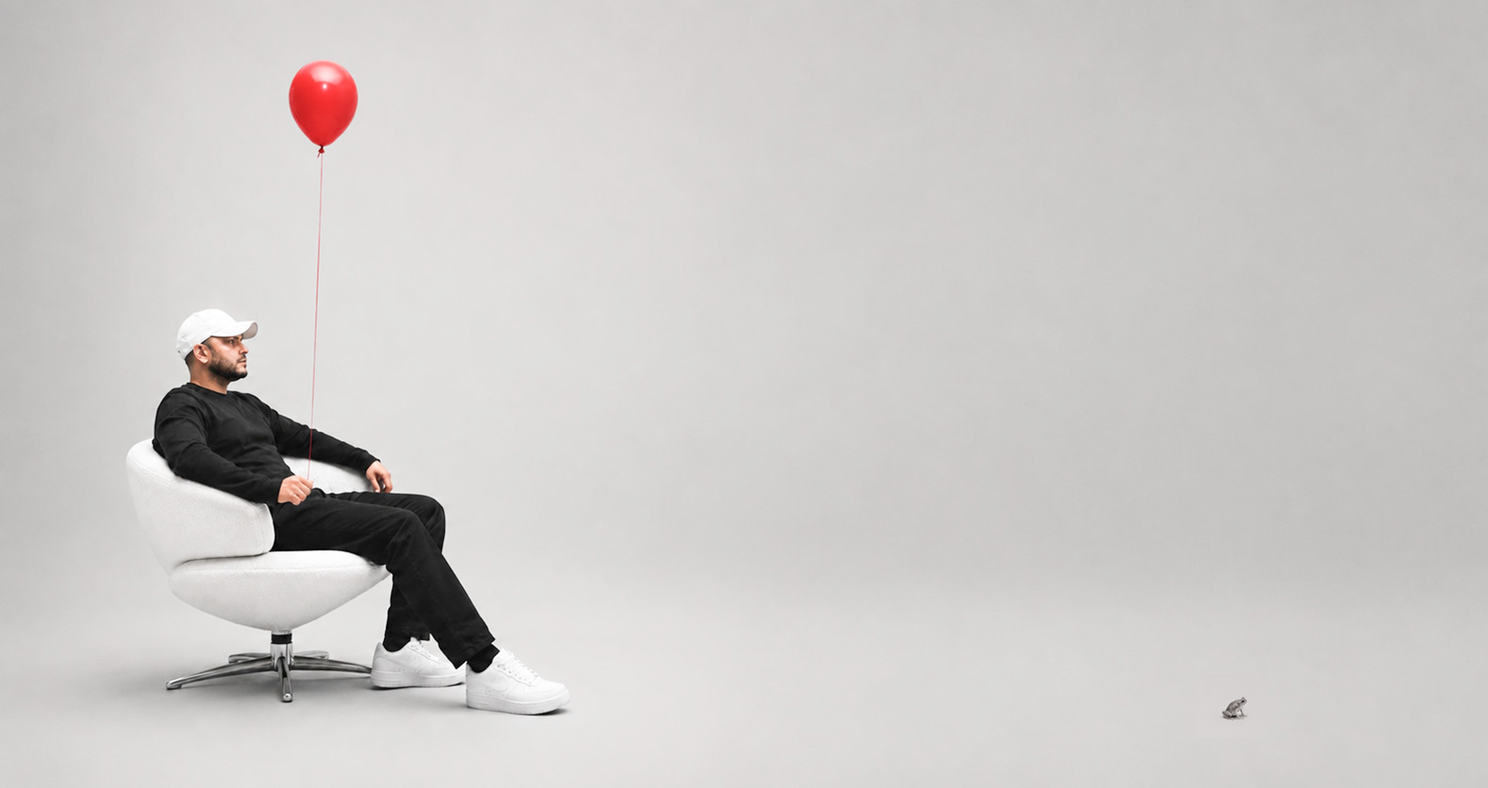 Nav Chatterji seated in a white chair holding a red balloon, with a small frog in the studio.